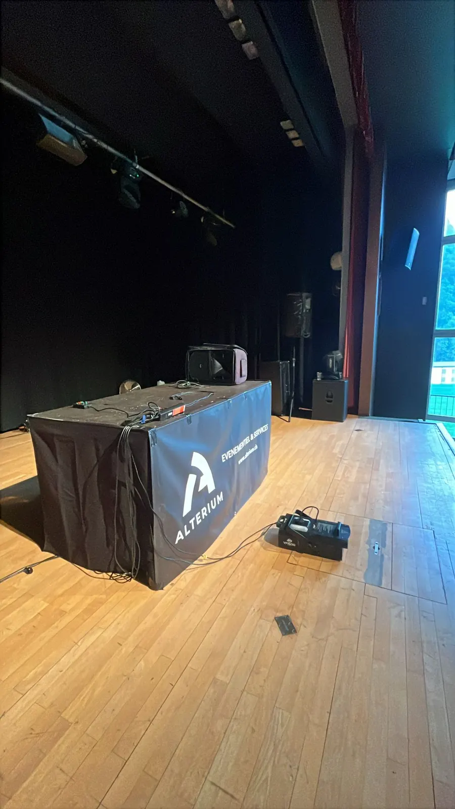 Régie DJ habillée d'une bâche Alterium sur la scène vide pendant l'installation, avec une machine à fumée au sol, une enceinte sur pied et une lyre posée sur un caisson de basse