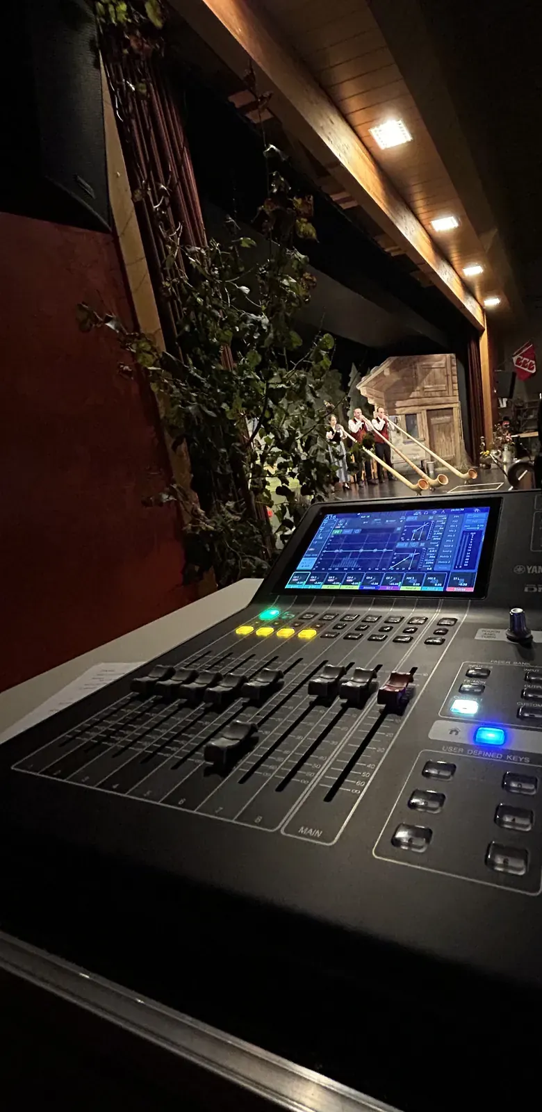 Console de mixage en régie à la salle de La Marelle, face à la scène où trois joueurs de cor des Alpes jouent devant un décor de chalet