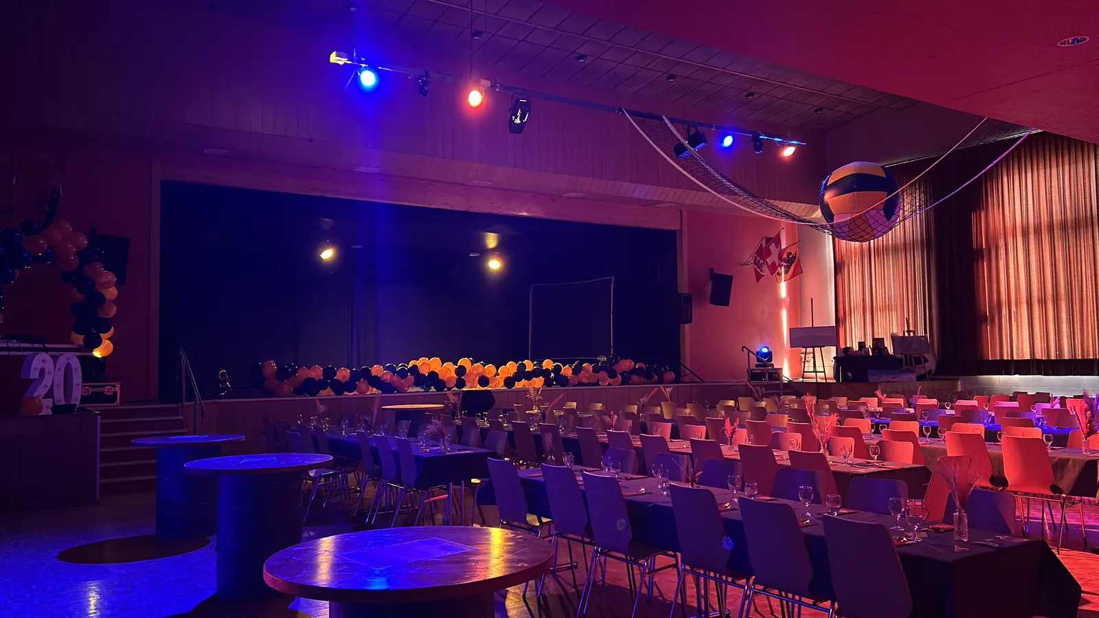 Salle dressée pour le souper des 20 ans du VBC Tramelan : tables en longues rangées, scène décorée de ballons orange et noirs, projecteurs sur une barre suspendue et lumière bleue et rouge